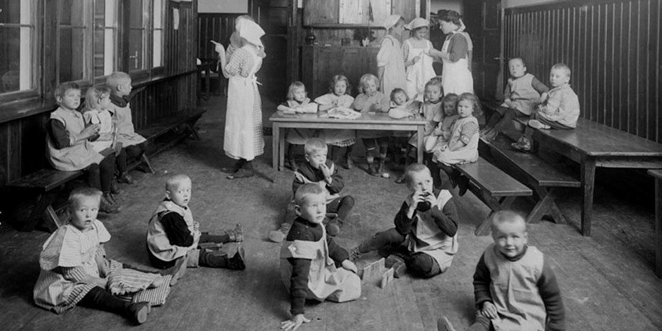 Vuxna och barn sitter på stolar och golv i ett rum tidigt 1900-tal. Foto.