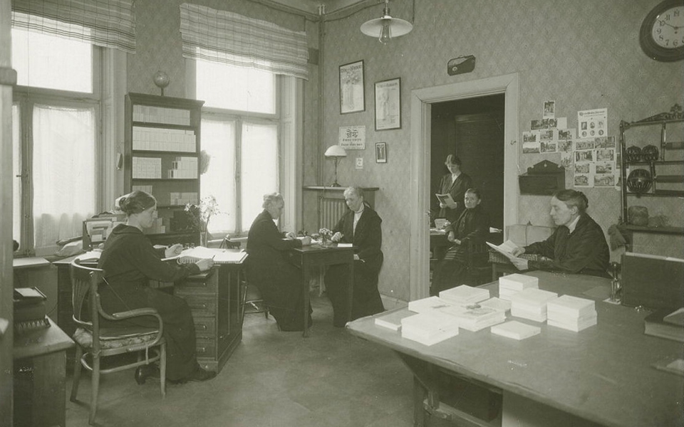 Några personer vid skrivbord och i ett rum i en publik lokal med fattigvård. Foto.